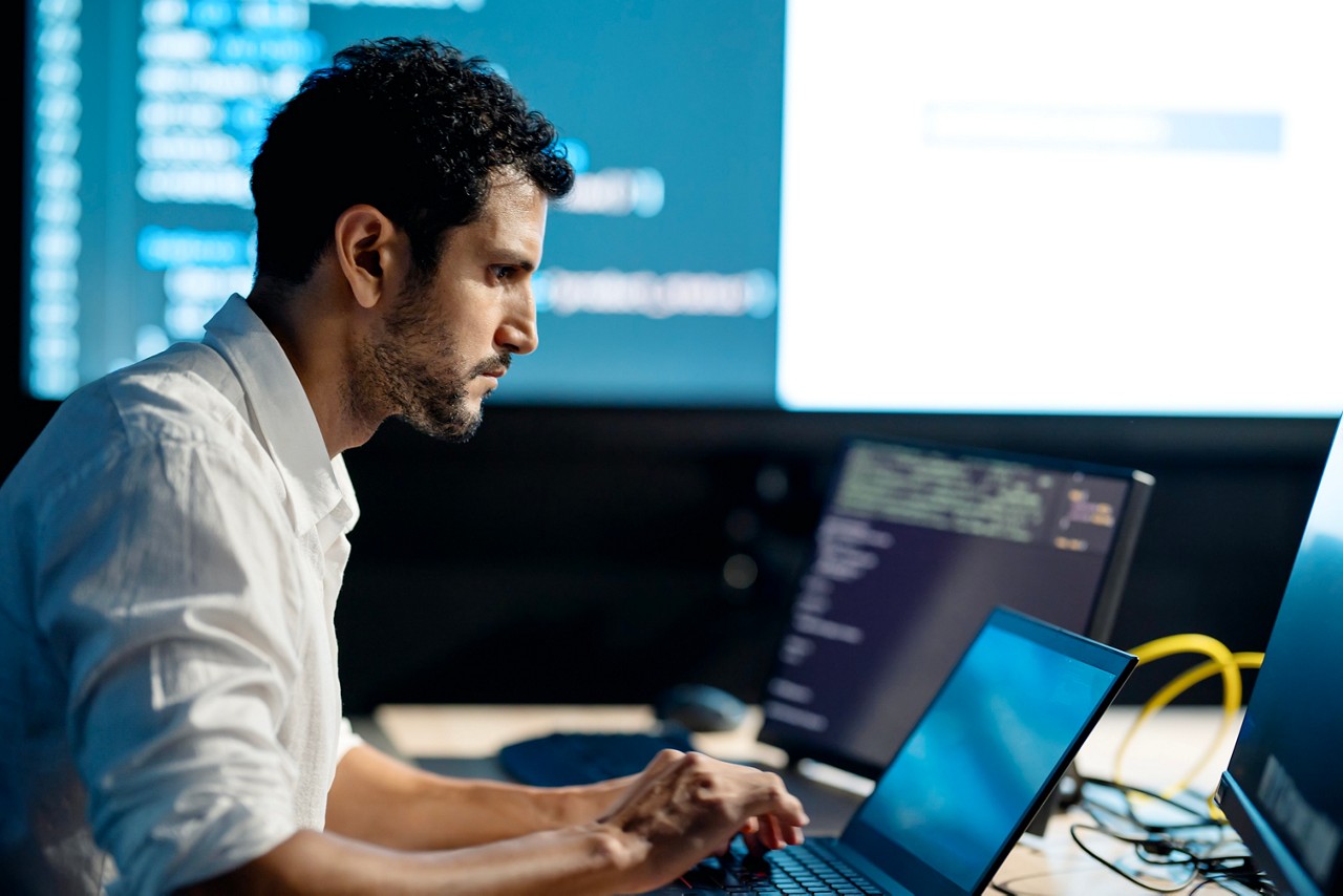 A man is seated at a desk working on a laptop. Multiple monitors display code and data in a dark room. The setting suggests a late-night tech session with intense focus on the screen.